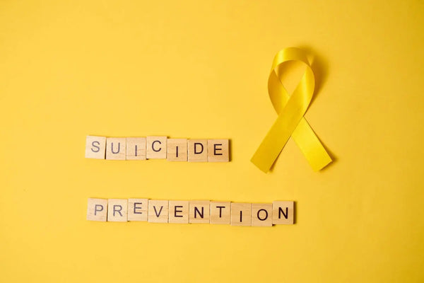 Gelbe Schleife und Holzbuchstaben mit der Aufschrift Suicide Prevention vor einem gelben Hintergrund.