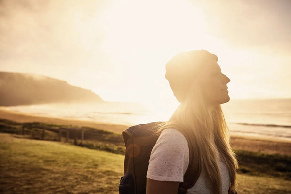 Junge Frau steht entspannt im Sonnenuntergang am Meer, atmet tief durch und wirkt gelassen, symbolisiert Achtsamkeit im Sommer