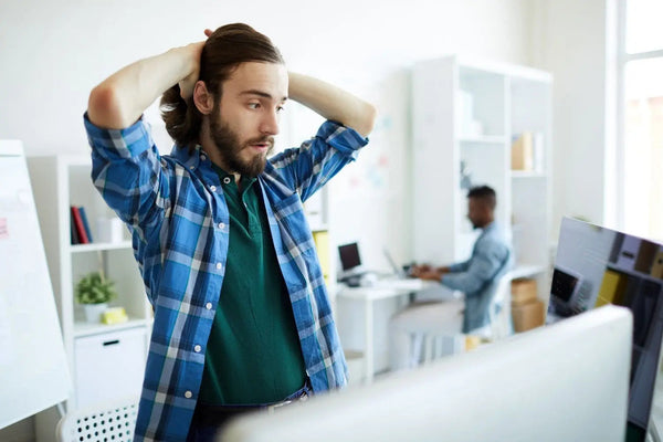 Junger Mann mit langem Haar im Büro, der sichtlich gestresst die Hände hinter den Kopf legt und angestrengt auf einen Computerbildschirm blickt.
